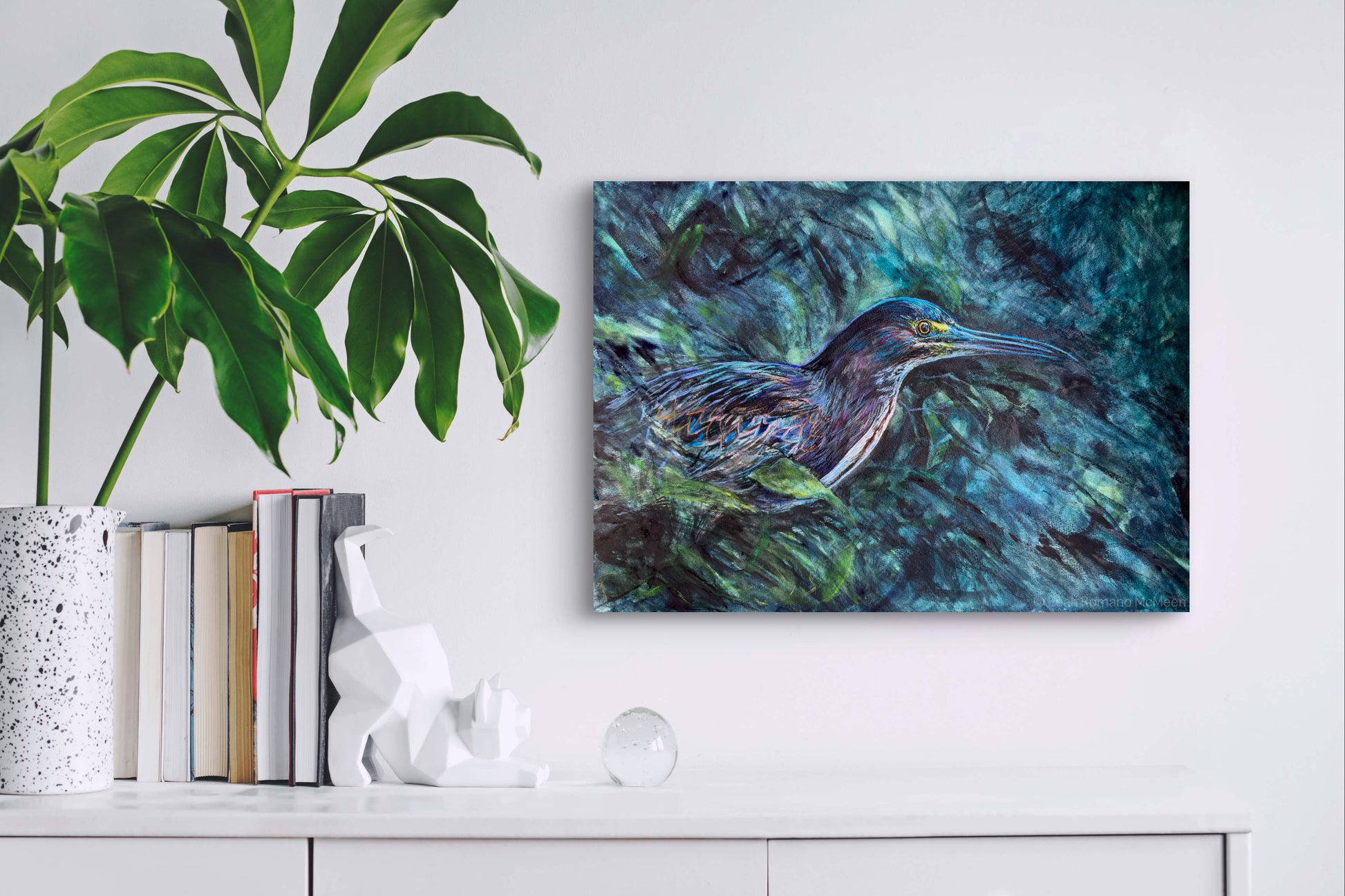 Framed artwork of a bird on a wall above a white cabinet with books and decor items.