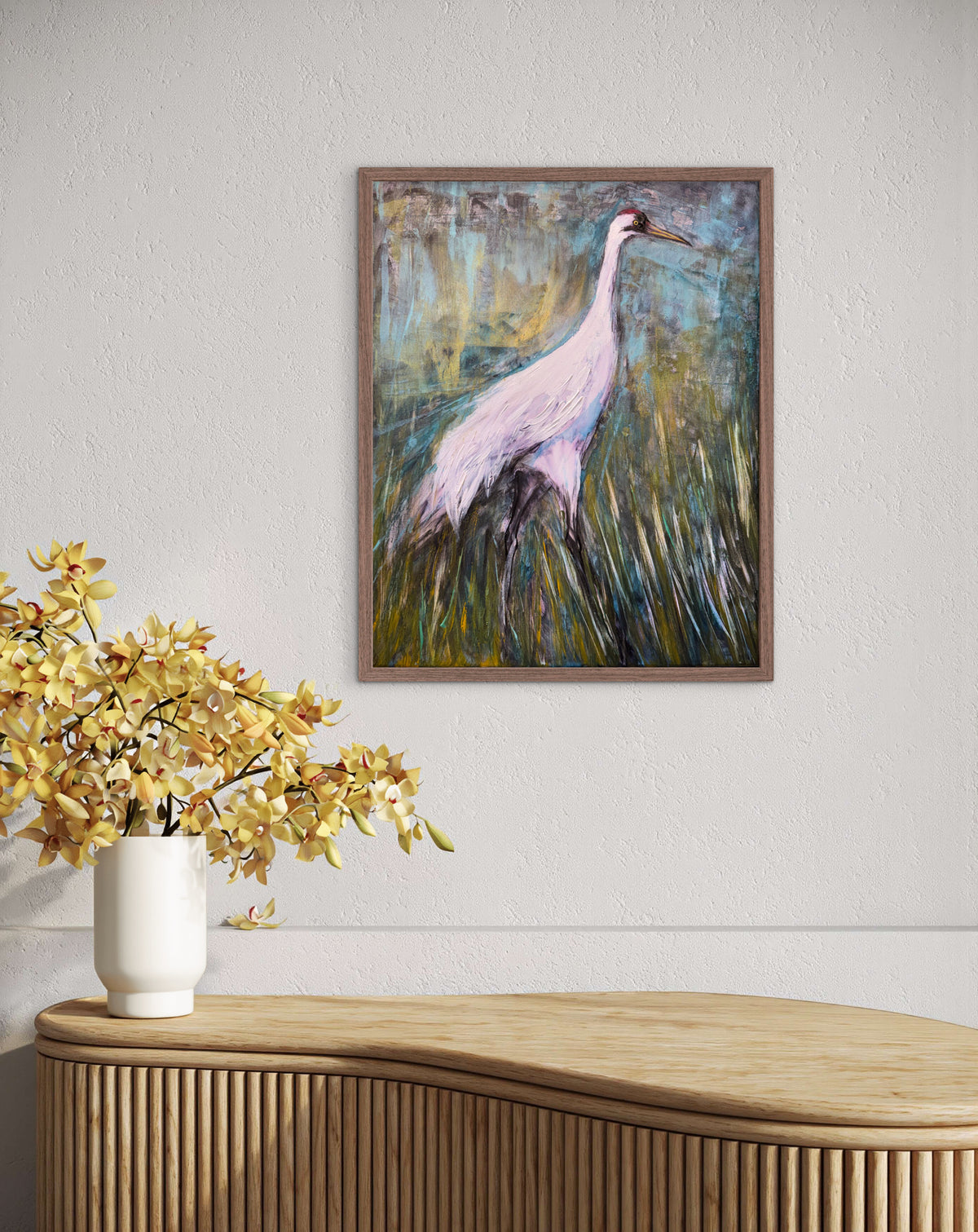 A framed painting of a white crane standing in tall grass hangs on a textured white wall above a light wood console table with a white vase of yellow flowers.