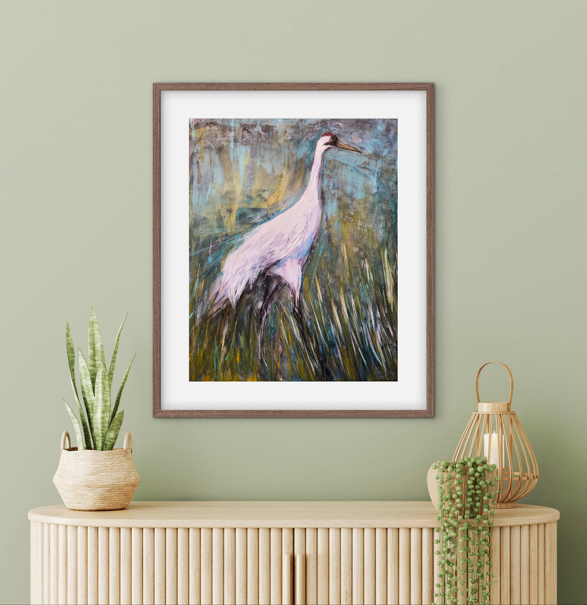 A framed art print of a white heron in a grassy field, displayed on a sage green wall above a light wood console table with decorative plants and a lantern.