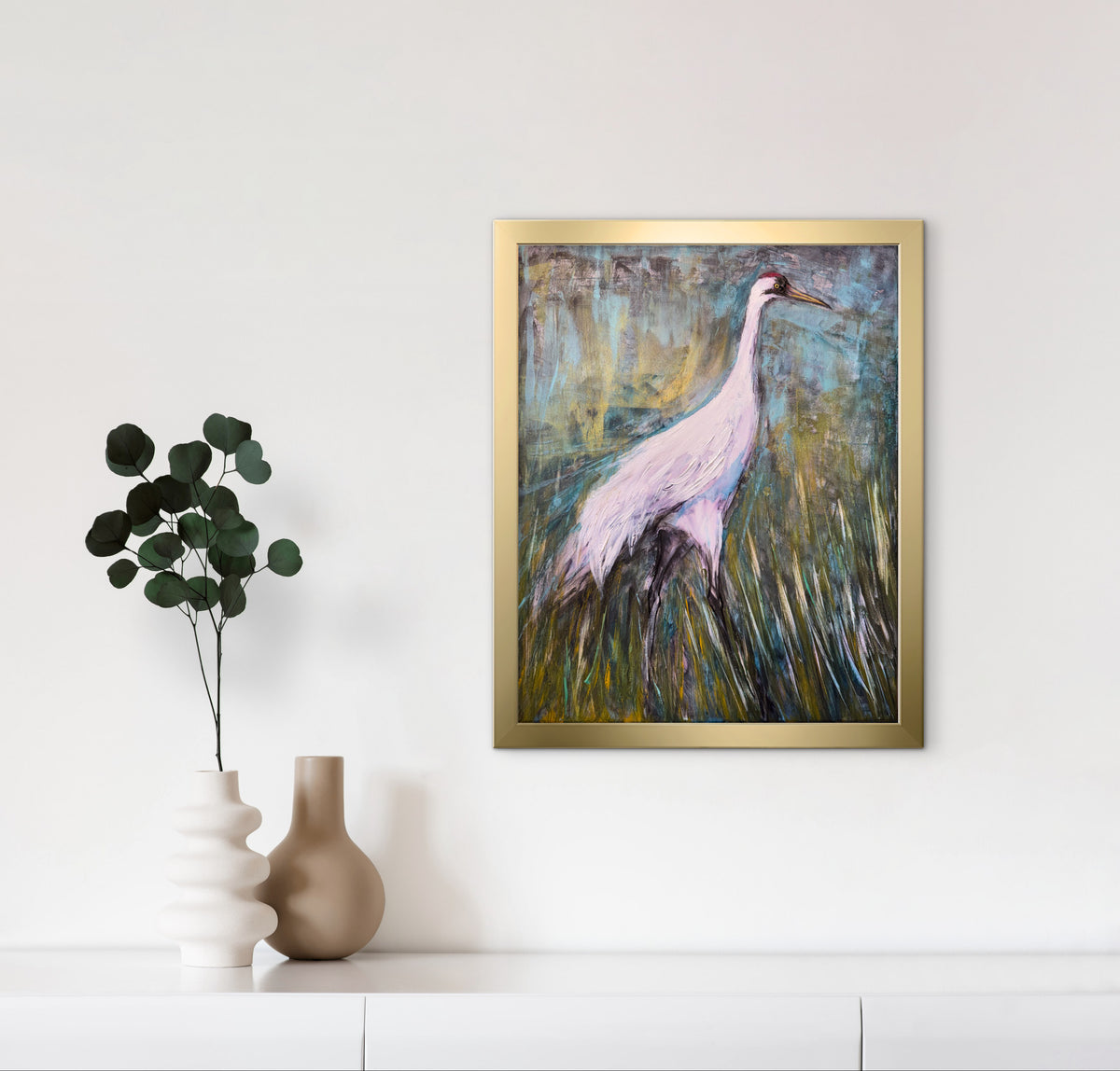 A framed painting of a pink heron standing in a grassy marsh, displayed on a white wall above a shelf with two vases.