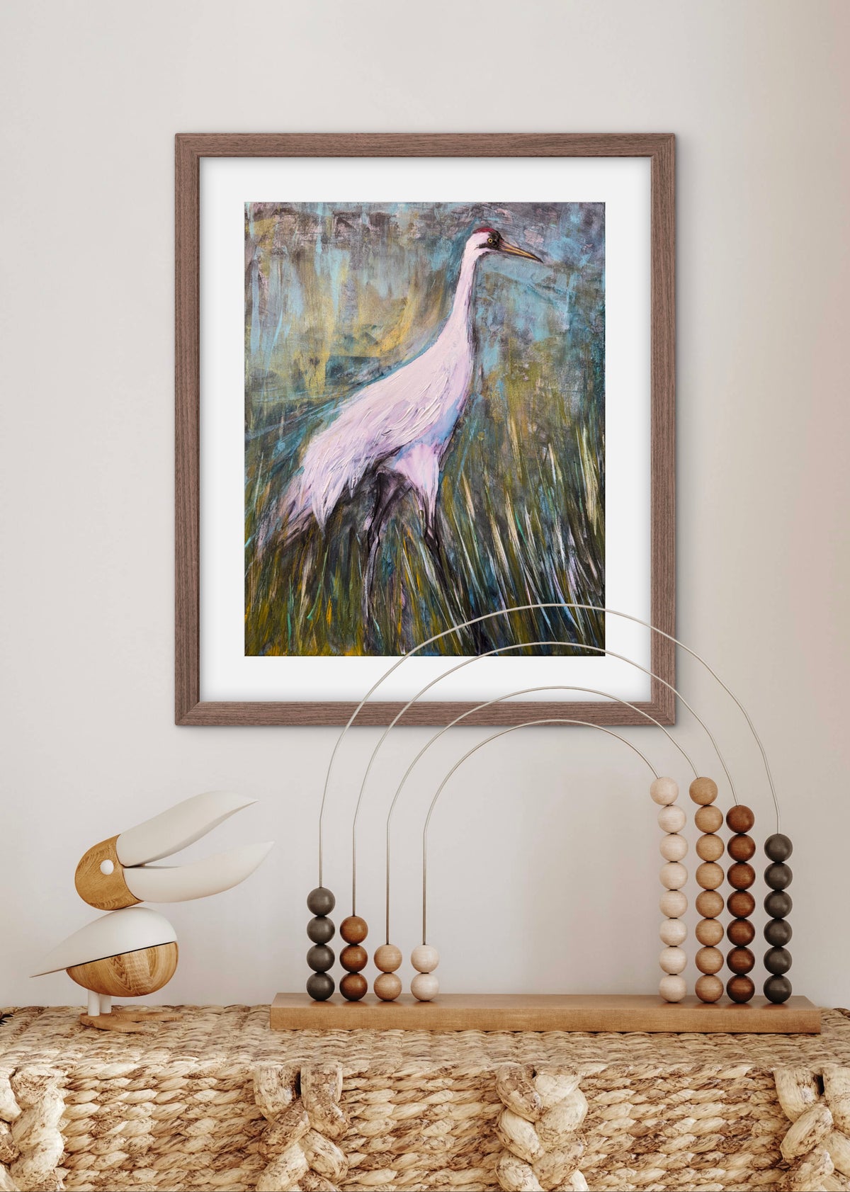 A framed art print of a heron in a marsh, styled on a woven table with decorative abacus-like objects against a neutral wall.
