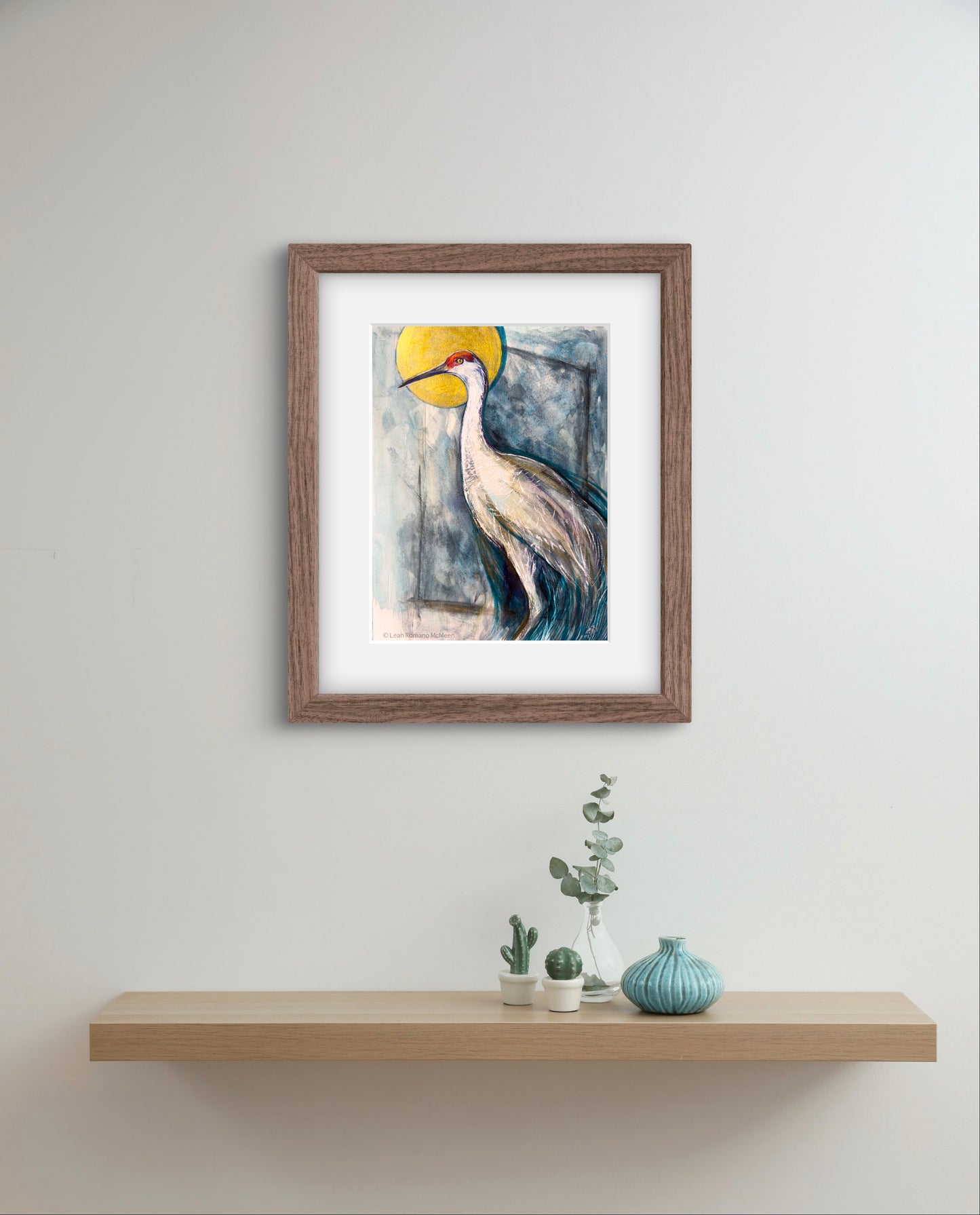 Framed artwork of a crane with a yellow sun on a white wall above a wooden shelf with decorative items.
