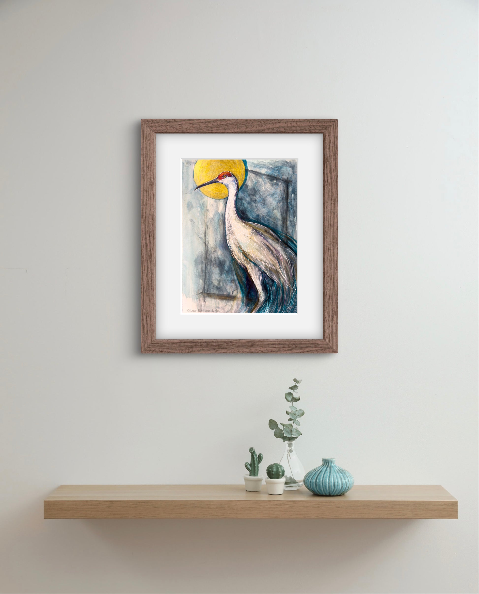 Framed artwork of a crane with a yellow sun on a white wall above a wooden shelf with decorative items.