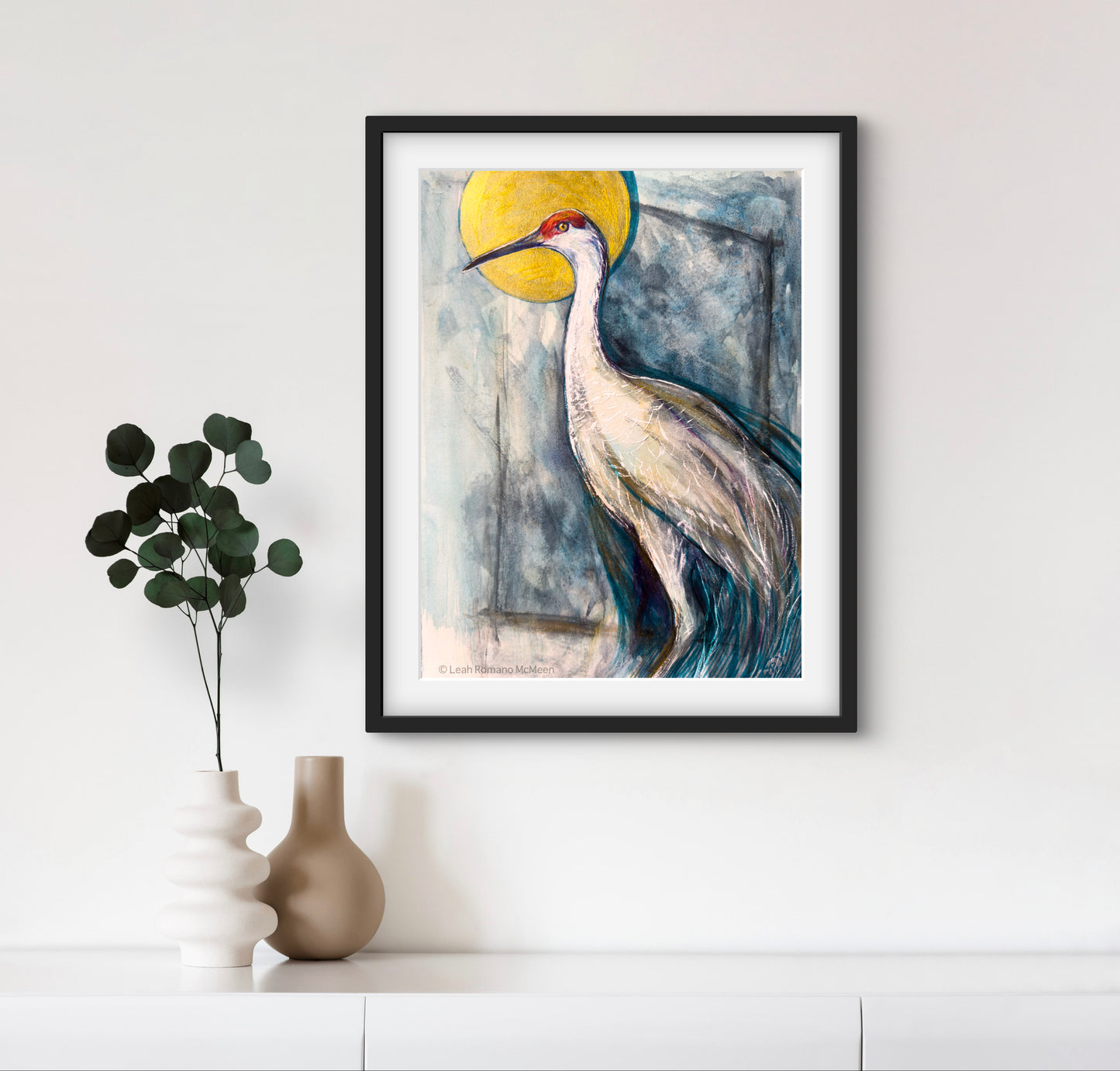 Framed artwork of a crane bird with a yellow sun on a white wall.
