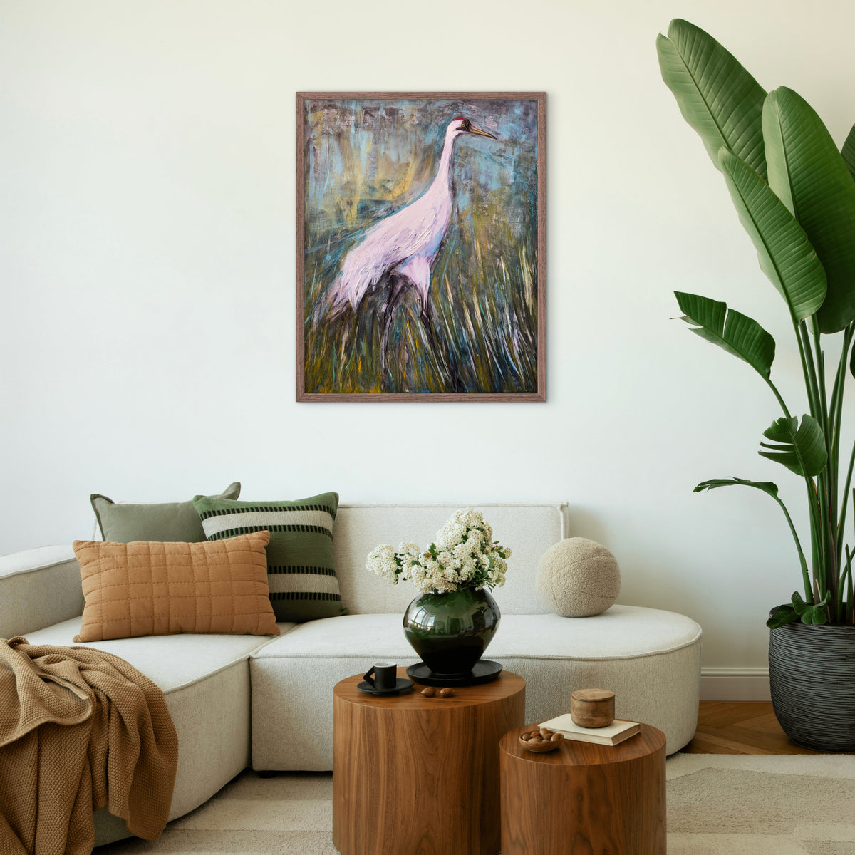 A framed painting of a pink flamingo in a grassy marsh hangs on a white wall above a modern beige sectional sofa in a styled living room with wooden side tables, a green vase of flowers, and a large potted plant.